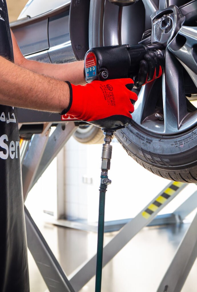 Technician wearing red gloves using an air-powered impact wrench on a car wheel while the vehicle is on a hydraulic lift.