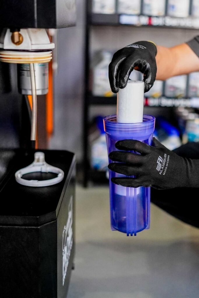 Worker wearing black gloves inserts a white filter into a blue reusable bottle under a beverage dispenser.